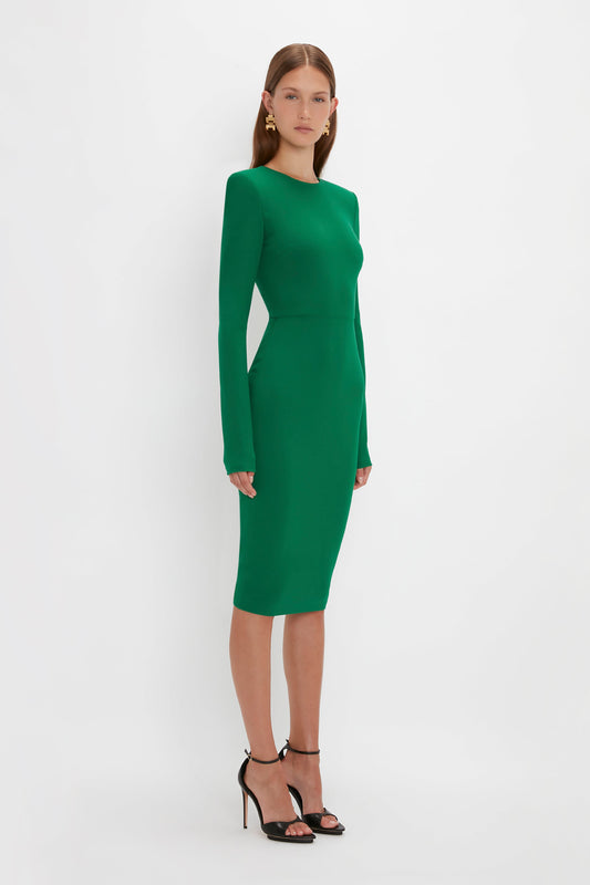 A woman stands in a Victoria Beckham Long Sleeve T-Shirt Fitted Dress in Emerald with padded shoulders and black high-heeled sandals against a white background.