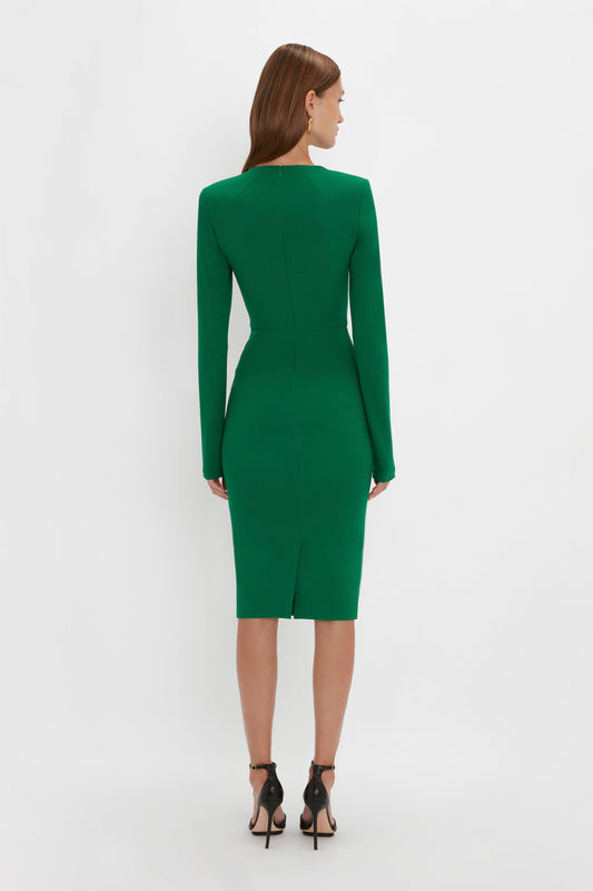 A woman with brown hair stands facing away, wearing the Long Sleeve T-Shirt Fitted Dress in Emerald by Victoria Beckham and black heels.
