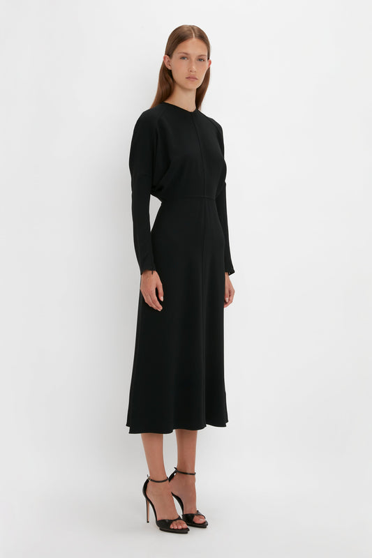 A woman in a simple Victoria Beckham Dolman Midi Dress in Black and black squared toe sandals stands against a white background.