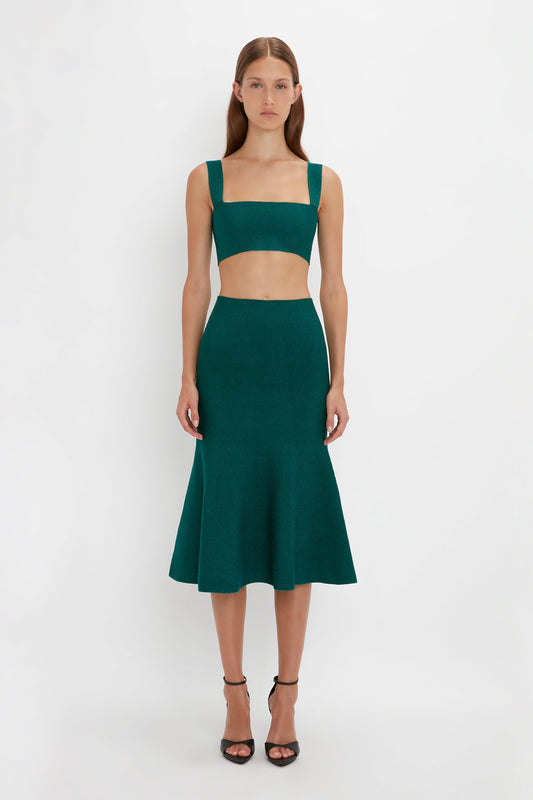 A woman stands wearing a green two-piece outfit featuring a VB Body Strap Bandeau Top In Lurex Green by Victoria Beckham and a VB Body Flared Skirt, paired with Pointy Toe Stiletto Sandals. She poses against a plain white background.
