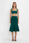 A woman stands against a plain white background, wearing a Lurex Green sleeveless crop top and a matching Victoria Beckham VB Body Flared Skirt In Lurex Green made from compact knitwear, paired with black strappy heels.