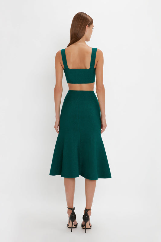 A woman with long hair stands facing away, wearing a sleeveless, green VB Body Strap Bandeau Top In Lurex Green by Victoria Beckham and black Pointy Toe Stiletto Sandals.