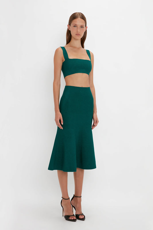 A woman with long hair wears a Victoria Beckham VB Body Strap Bandeau Top In Lurex Green and a VB Body flared skirt in teal that flares below the knees. She stands against a plain white background, wearing pointy toe stiletto sandals.