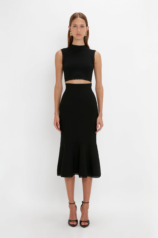 A person is standing against a plain white background, wearing a versatile VB Body Scallop Trim Tank Top In Black from Victoria Beckham with a cutout at the midsection and a flared hemline, paired with black heels.