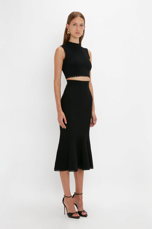 A woman stands against a plain white background wearing a versatile black Victoria Beckham VB Body Scallop Trim Tank Top In Black paired with a matching high-waisted black midi skirt with a flared hem. She completes her summer essential look with black high-heeled sandals.