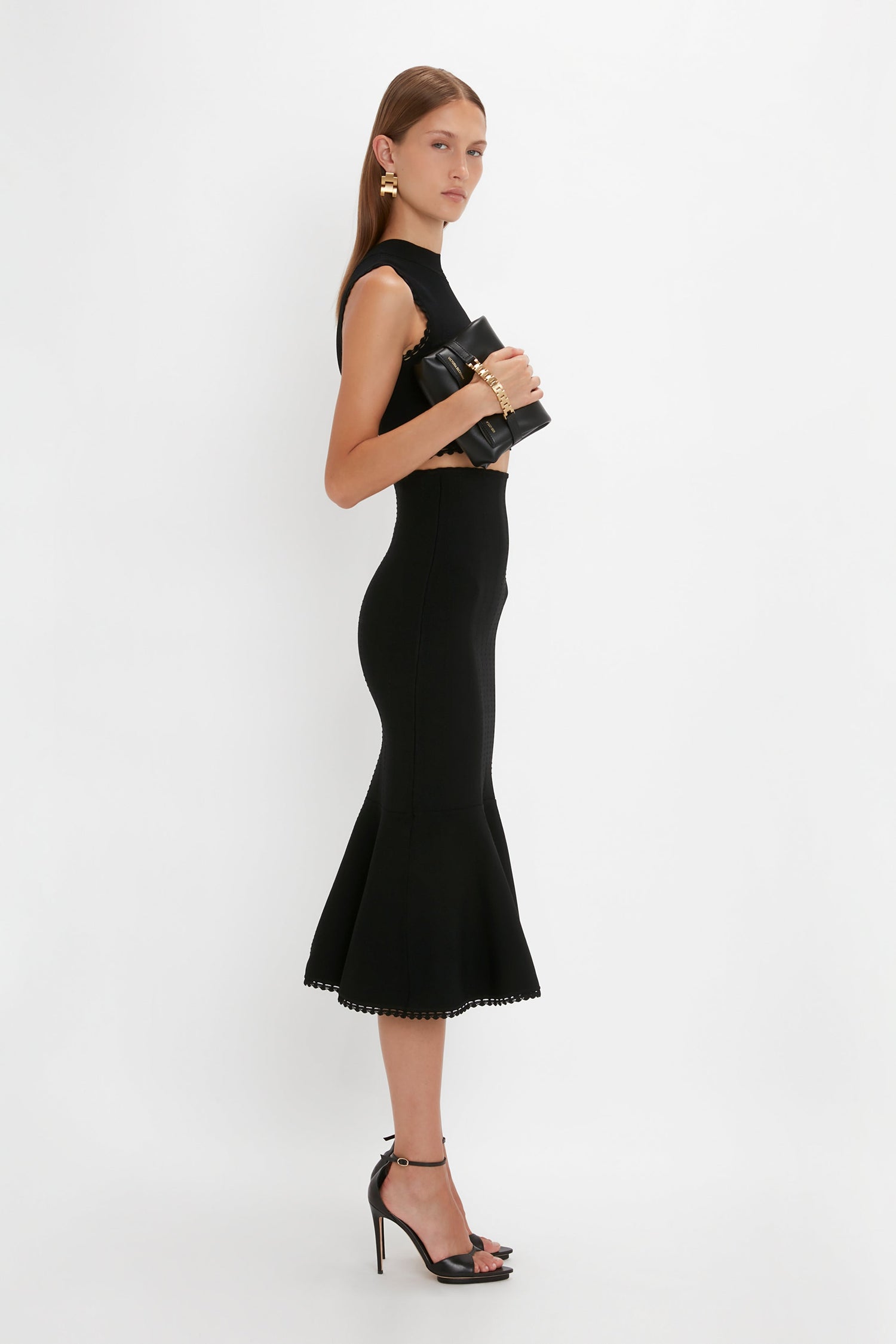 A person stands sideways wearing a sleeveless Victoria Beckham VB Body Scallop Trim Flared Skirt In Black with a flared silhouette and scallop trim, paired with black high-heeled sandals, and holding a black clutch bag.