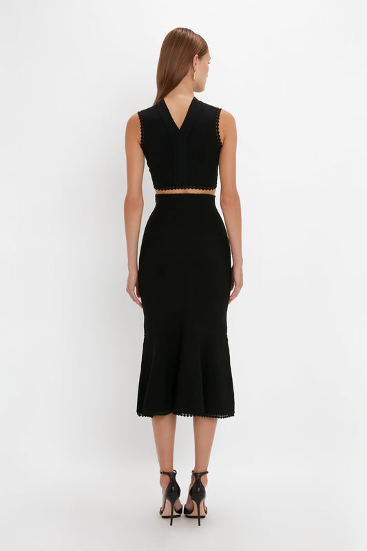 A woman with long hair is standing with her back to the camera, wearing a versatile Victoria Beckham VB Body Scallop Trim Tank Top In Black and a matching high-waisted, fitted black skirt paired with black high-heeled sandals—an elegant summer essential.