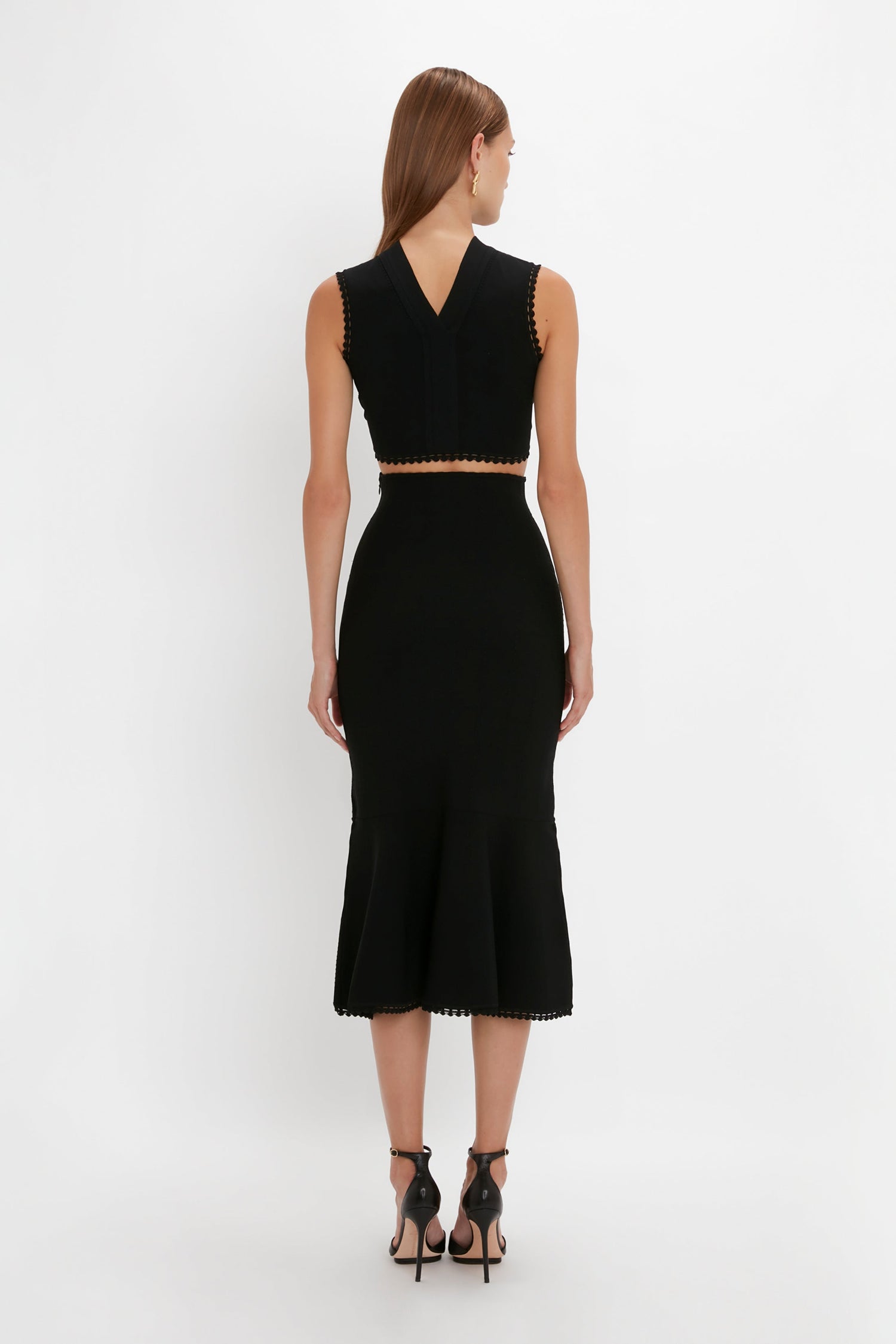 A person with long hair is shown from the back wearing a black, sleeveless, V-neck crop top paired with a high-waisted VB Body Scallop Trim Flared Skirt In Black by Victoria Beckham. They complete the look with black high-heeled shoes.