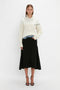 A woman stands against a plain white background, wearing a Collar Detail Jumper In Natural by Victoria Beckham that exudes casual sophistication. Paired with a black midi skirt and black heeled sandals, she completes her look with one hand in her skirt pocket, effortlessly blending style and ease.