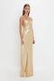 Person standing wearing a long, 1990s-inspired Victoria Beckham Exclusive Floor-Length Cami Dress In Gold with spaghetti straps, set against a plain white background.