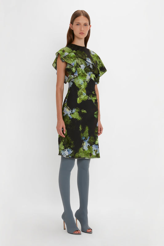 A woman stands against a white background wearing a black Twist Shoulder Dress In Black Frost by Victoria Beckham with green and blue floral patterns and gray open-toe thigh-high boots. The dress features an asymmetric hemline, complementing her straight brown hair and neutral expression.