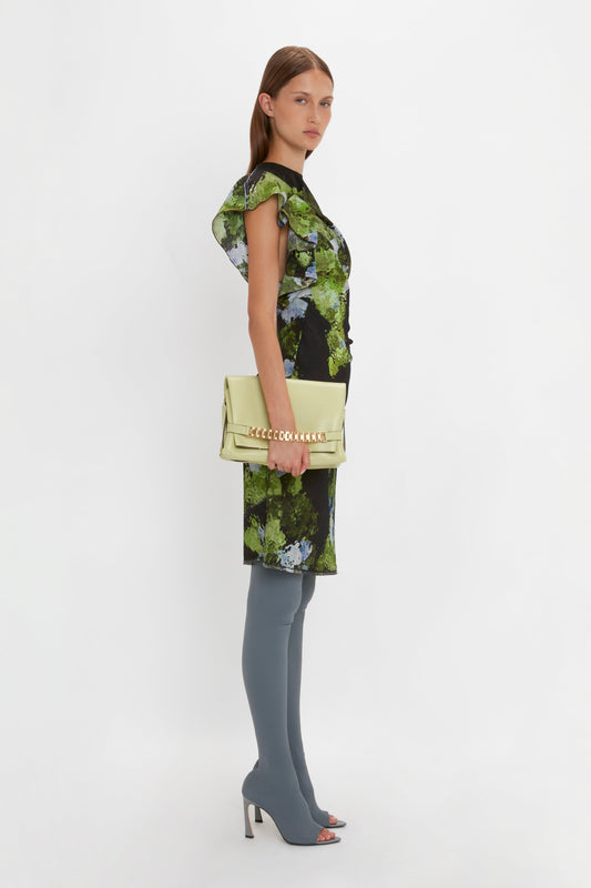 A person with long hair is dressed in a green and black floral Twist Shoulder Dress In Black Frost by Victoria Beckham with draped pleat details, paired with gray over-the-knee boots. They are holding a cream-colored clutch and standing sideways against a white background.