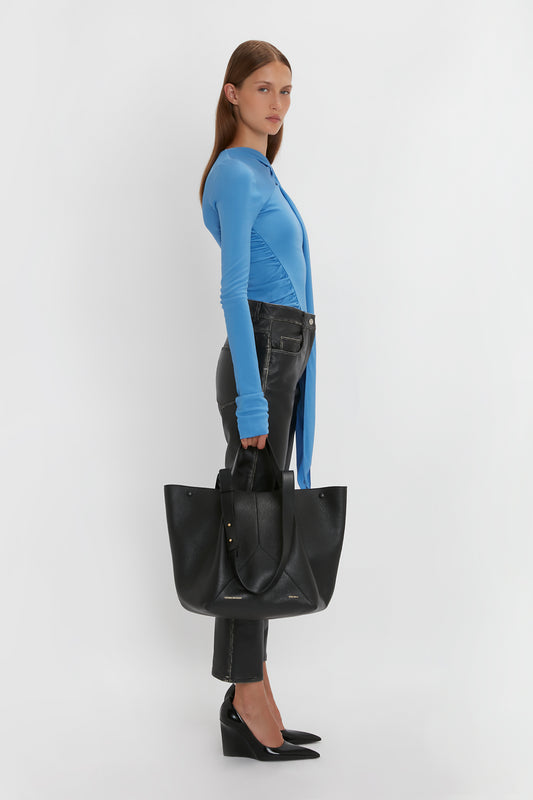 A person wearing a long-sleeve blue top, black pants, and black shoes is holding a large W11 Medium Tote Bag In Black Leather by Victoria Beckham while looking to the side.