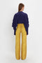 A person with long hair stands facing away, wearing a Tie Detail Ruffle Blouse In Ultraviolet by Victoria Beckham with a V neckline and yellow patterned wide-leg pants.