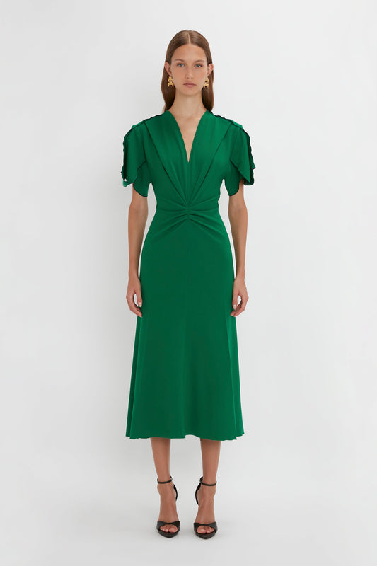 A woman in a vibrant green Gathered V-Neck Midi Dress in Emerald by Victoria Beckham, with ruffled sleeves and a waist-defining pleat, standing against a plain white background.