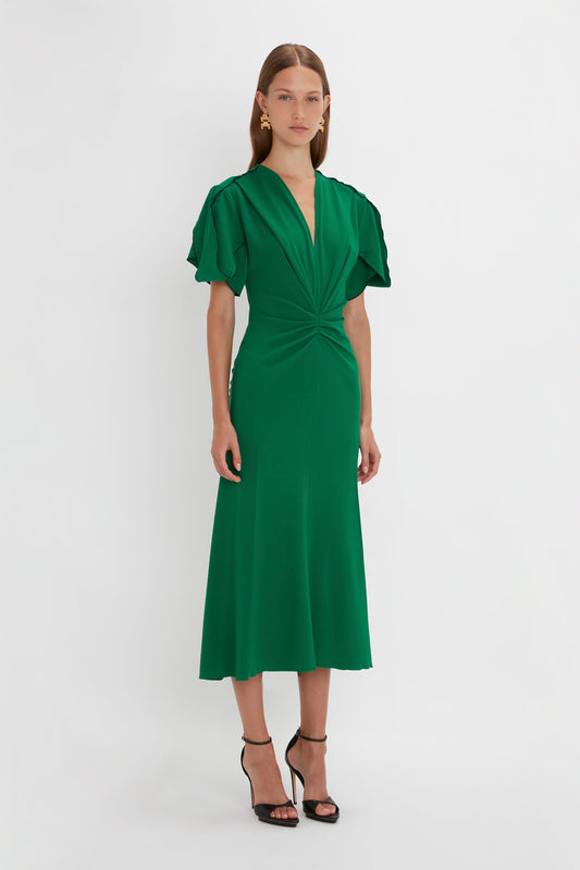 A woman in a Victoria Beckham Gathered V-Neck Midi Dress in Emerald with ruffled sleeves and a waist-defining pleat, standing against a white background, wearing black heels.
