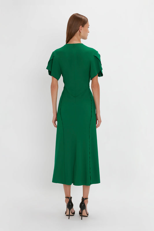 A woman in a Victoria Beckham Gathered V-neck Midi Dress in Emerald with ruffle sleeves and a slit, paired with black heels, viewed from the back against a white background.