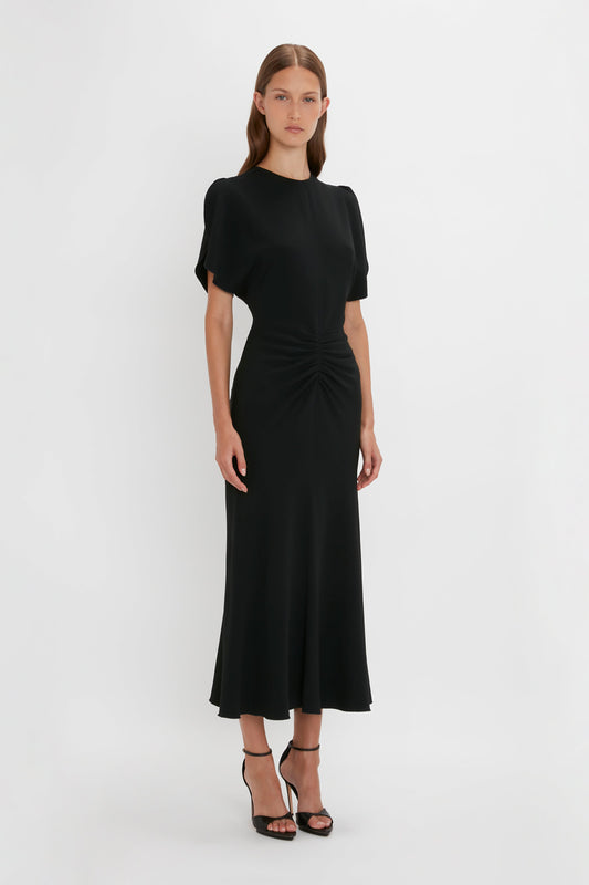A woman in a black Victoria Beckham Gathered Waist Midi Dress with short sleeves and ruched detail at the waist, standing against a plain white background.