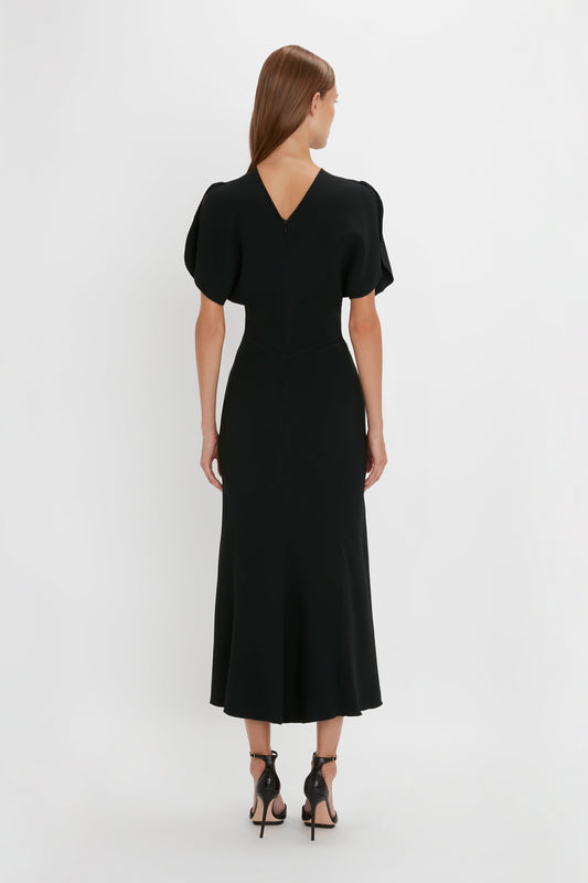Woman standing with her back to the camera, wearing a Victoria Beckham Gathered Waist Midi Dress in Black with short sleeves and v-back, paired with black pointy toe stilettos on a white backdrop.