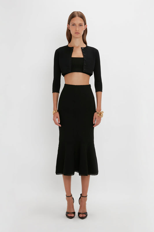 A person stands against a white background wearing a black crop top, a VB Body Cropped Cardigan In Black by Victoria Beckham, and a black midi skirt that features a form-fitting silhouette paired with black high-heeled sandals. They have minimal accessories.