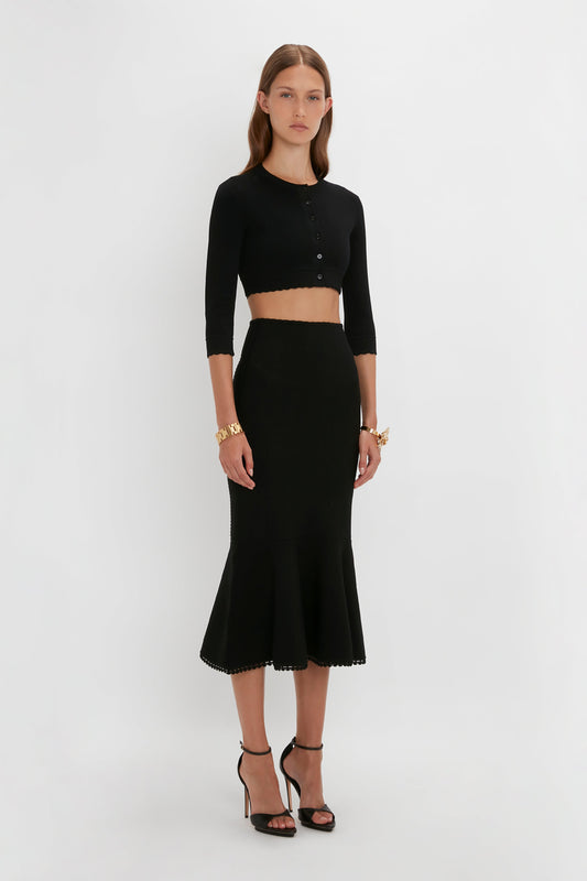 A woman wearing a black VB Body Cropped Cardigan In Black by Victoria Beckham and a high-waisted black midi skirt with a flared hem stands against a plain white background. The form-fitting silhouette is accessorized with bracelets and black high-heeled sandals, enhancing the ensemble's feminine detailing.