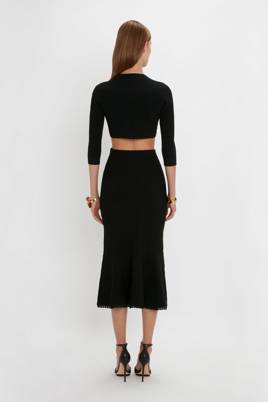 A woman stands facing away, wearing a black VB Body Cropped Cardigan In Black by Victoria Beckham and a matching black skirt with feminine detailing. She has long brown hair and is accessorized with gold bracelets and black high-heeled sandals, creating a form-fitting silhouette.