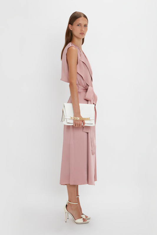 A person in a light pink sleeveless dress stands against a white background, holding a white Chain Pouch Bag with Strap In White Leather by Victoria Beckham and wearing white high-heeled shoes.