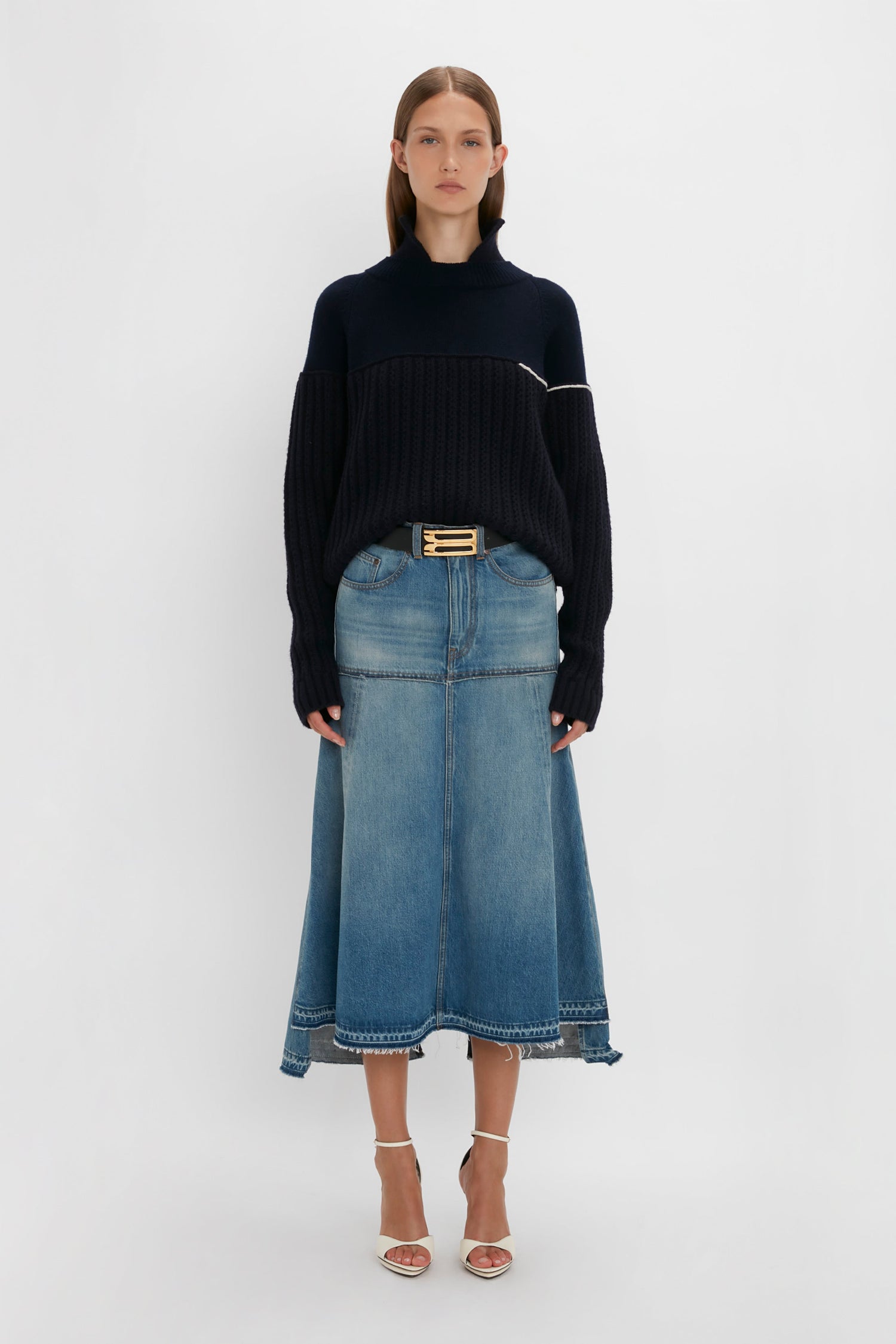 A woman stands wearing a chic, Collar Detail Jumper In Navy by Victoria Beckham paired with a knee-length blue denim skirt and white heeled sandals against a plain white background.