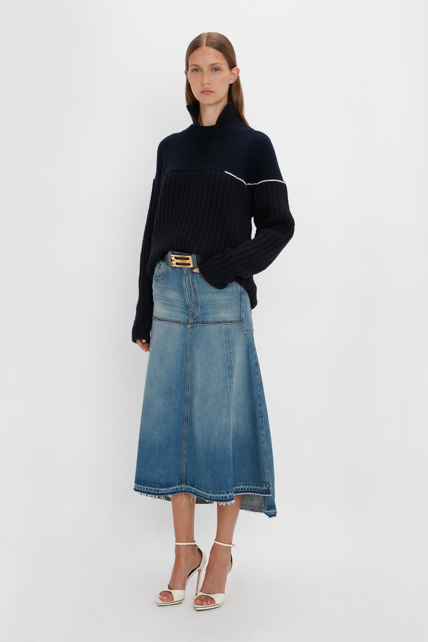 A woman stands against a plain background, wearing the Collar Detail Jumper In Navy by Victoria Beckham, a mid-length denim skirt, and white high-heeled sandals. She has straight, shoulder-length hair and one hand in her pocket.