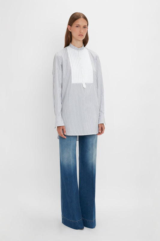 Person standing against a plain background, wearing the Tuxedo Bib Shirt in Black and Off-White by Victoria Beckham and wide-leg blue jeans, showcasing menswear silhouettes with a roomy fit.