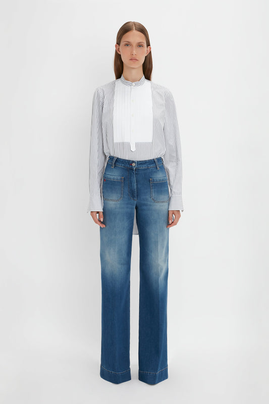 A person stands against a neutral background, wearing the Victoria Beckham Tuxedo Bib Shirt in Black and Off-White with vertical stripes and wide-leg blue jeans that boast a roomy fit.
