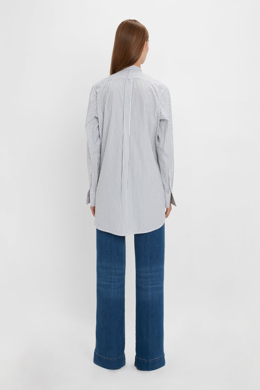 Person with long hair standing with their back to the camera, wearing a Tuxedo Bib Shirt in Black and Off-White by Victoria Beckham and blue denim jeans made from organic cotton, showcasing relaxed menswear silhouettes against a white background.