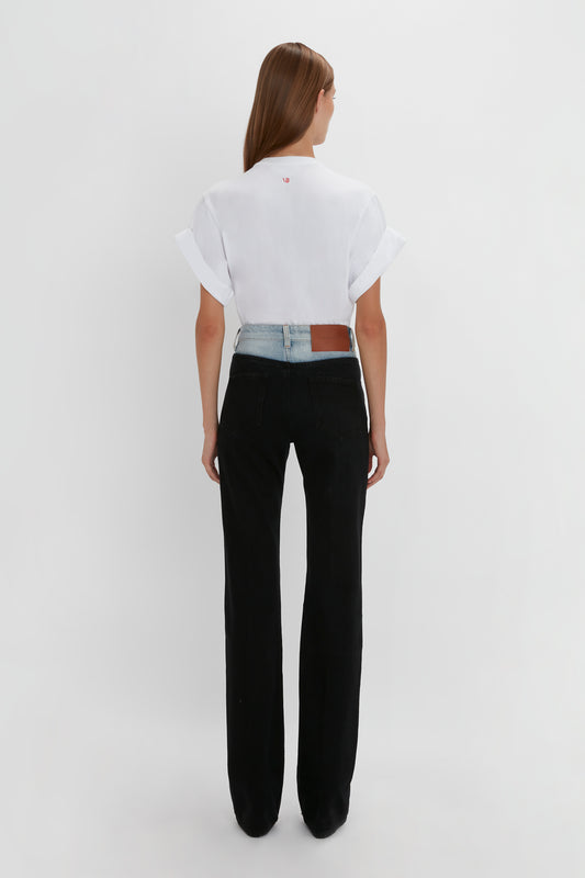 Woman facing away, wearing a white t-shirt and black Victoria Beckham Julia Jean In Contrast Wash high-waisted flared jeans, against a plain white background.