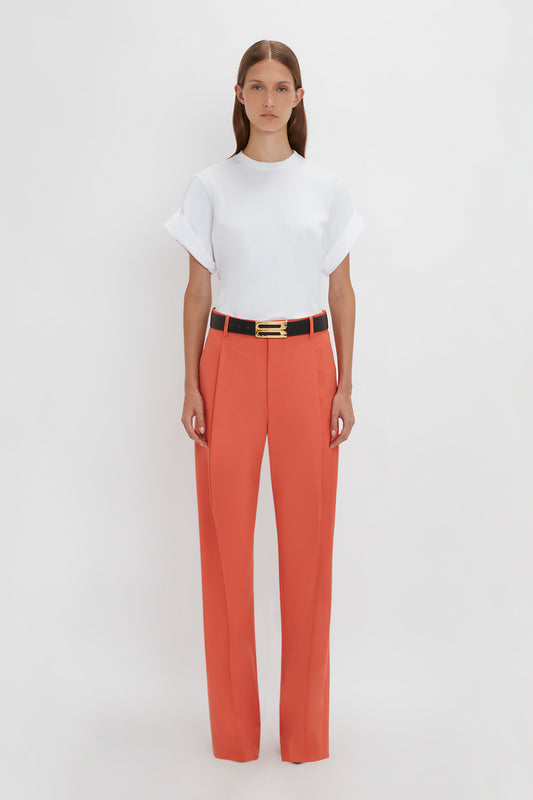A person standing against a plain white background, wearing a white short-sleeved shirt and Victoria Beckham Single Pleat Trouser In Papaya with wide legs, cinched with a black belt.