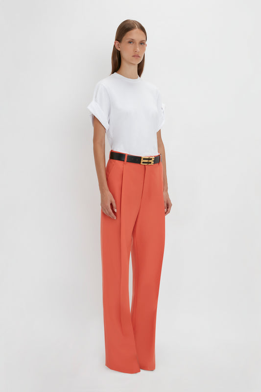 A person stands against a plain white background, wearing a white rolled-sleeve t-shirt and high-waisted Single Pleat Trouser In Papaya by Victoria Beckham with a black belt.