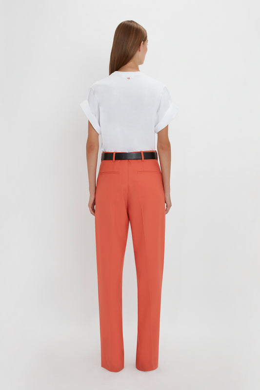 A person wearing an untucked white short-sleeve shirt and high-waisted, wide-leg Single Pleat Trouser In Papaya by Victoria Beckham with a black belt. The individual is standing facing away from the camera against a plain white background.