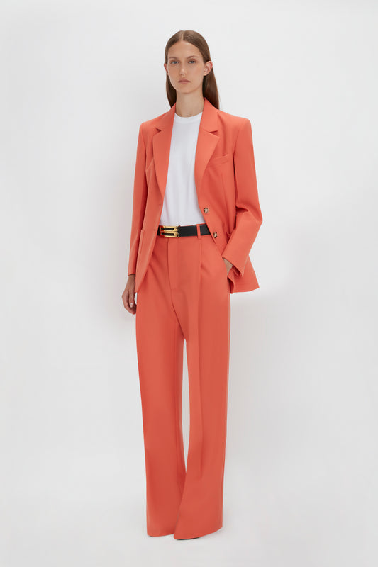A person stands against a plain white background wearing a coral-colored Victoria Beckham Single Pleat Trouser In Papaya with wide-legged pants, paired with a white T-shirt.