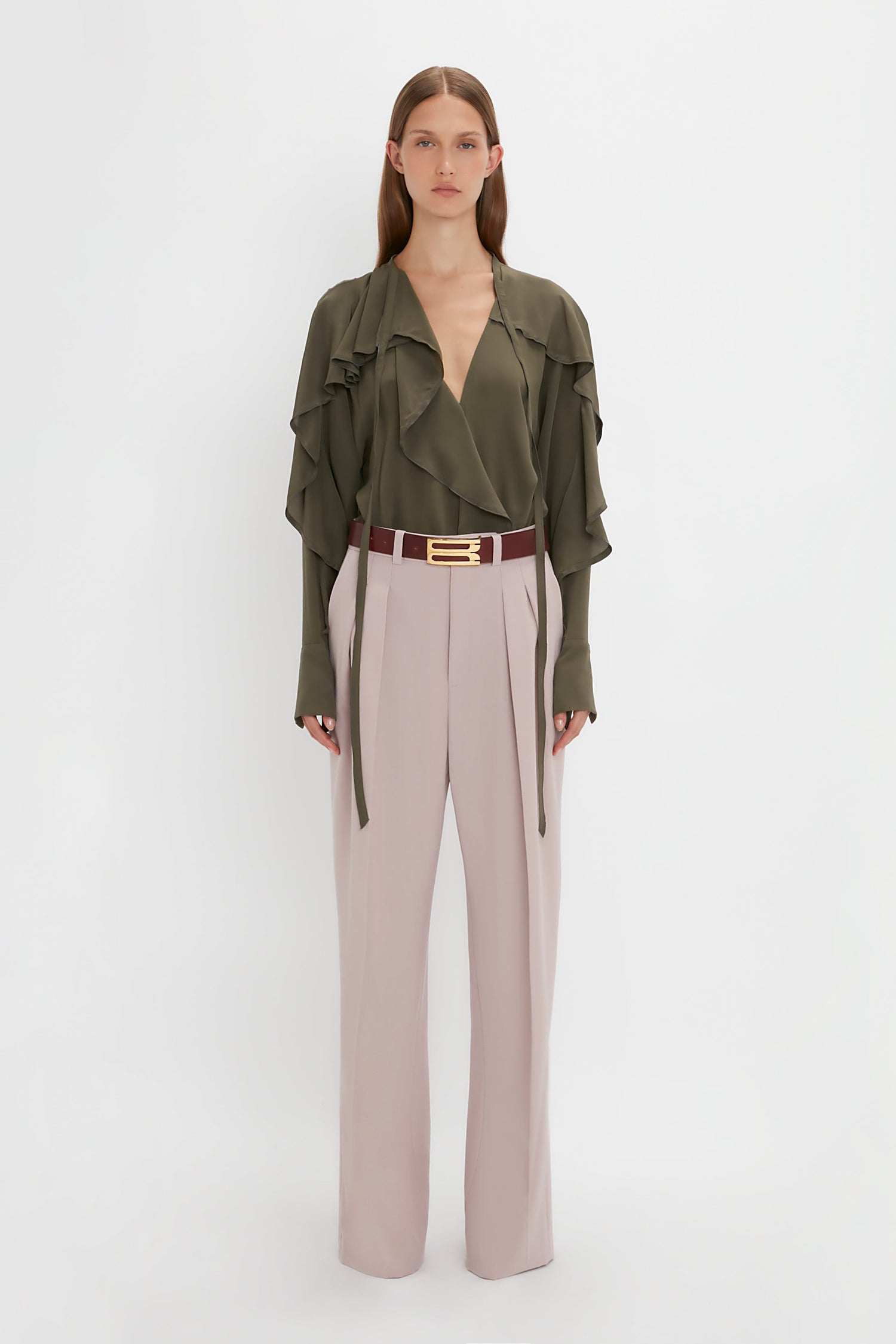 A woman with straight brown hair wears a green Tie Detail Ruffle Blouse in Oregano by Victoria Beckham, light pink wide-leg pants, and a maroon belt with a gold buckle, standing against a plain white background.