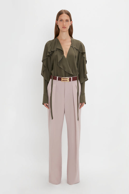 A person standing against a white background, wearing a green ruffled blouse, Victoria Beckham Double Pleat Trouser In Rose Quartz, and a maroon belt.