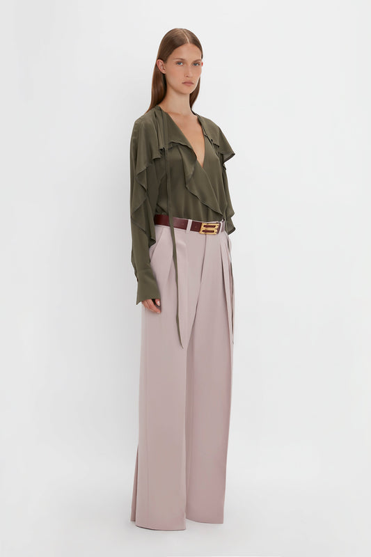 A woman stands against a plain white background wearing a green ruffled blouse tucked into high-waisted, wide-leg Double Pleat Trouser In Rose Quartz by Victoria Beckham, secured with a brown belt, showcasing a directional silhouette.