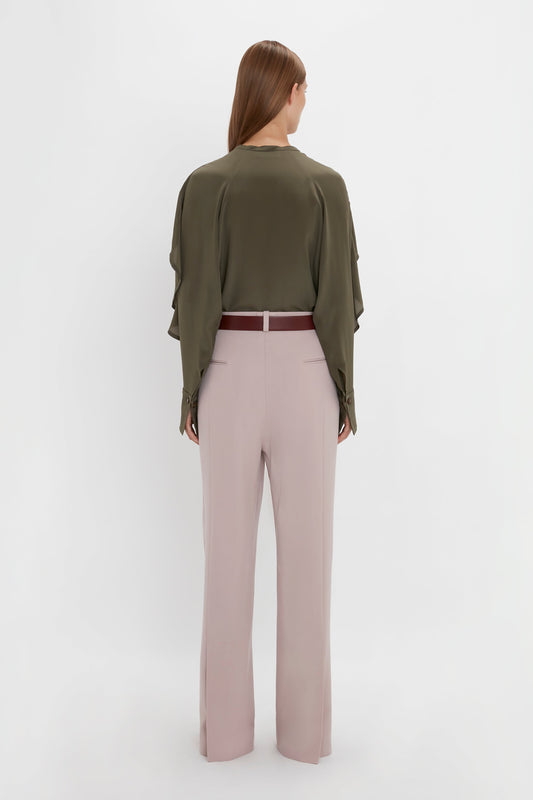 A person with long hair stands facing away, their directional silhouette accentuated by a dark green blouse and Victoria Beckham's Double Pleat Trouser In Rose Quartz with a red belt, set against a plain white background.