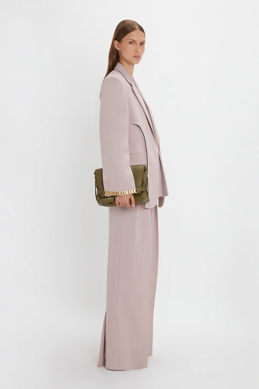 A person with long, straight hair wearing a light pink suit and a Double Panel Front Jacket In Rose Quartz by Victoria Beckham holds a green handbag with a gold chain. They stand against a plain white background, facing slightly to the right.