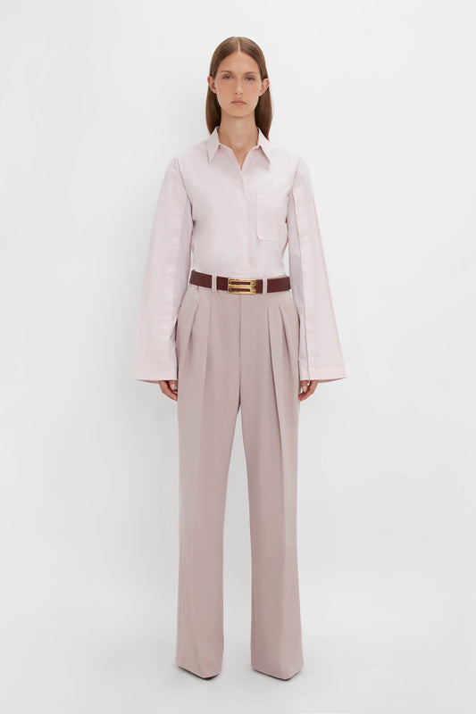 A person stands against a white background, wearing a Button Detail Cropped Shirt In Rose Quartz by Victoria Beckham, tucked into high-waisted beige trousers with pleats, secured with a brown belt.