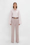 A person stands against a white background, wearing a Button Detail Cropped Shirt In Rose Quartz by Victoria Beckham, tucked into high-waisted beige trousers with pleats, secured with a brown belt.