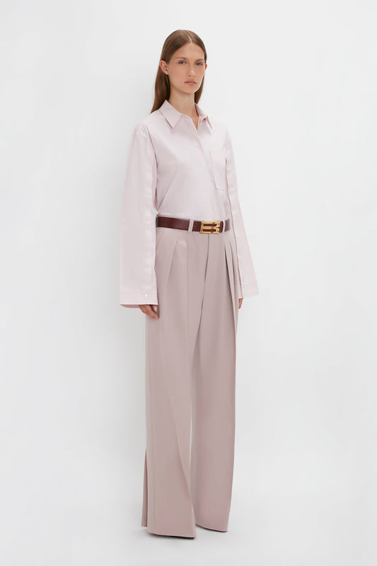 A woman stands against a plain white background wearing a Victoria Beckham Button Detail Cropped Shirt in Rose Quartz, beige wide-leg trousers, and a brown belt.