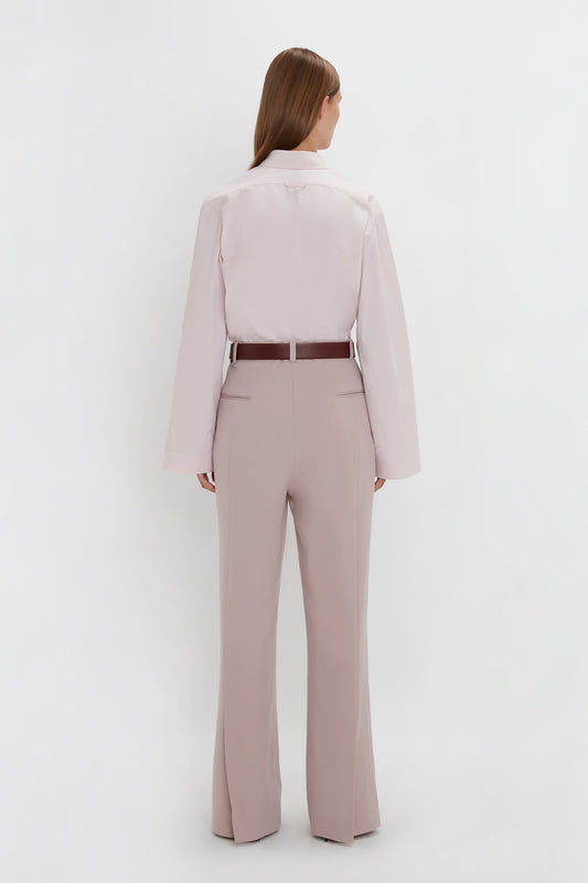 A person with long hair is standing with their back to the camera, wearing a Button Detail Cropped Shirt In Rose Quartz by Victoria Beckham, tucked into high-waisted taupe trousers.
