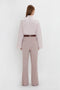 A person with long hair is standing with their back to the camera, wearing a Button Detail Cropped Shirt In Rose Quartz by Victoria Beckham, tucked into high-waisted taupe trousers.