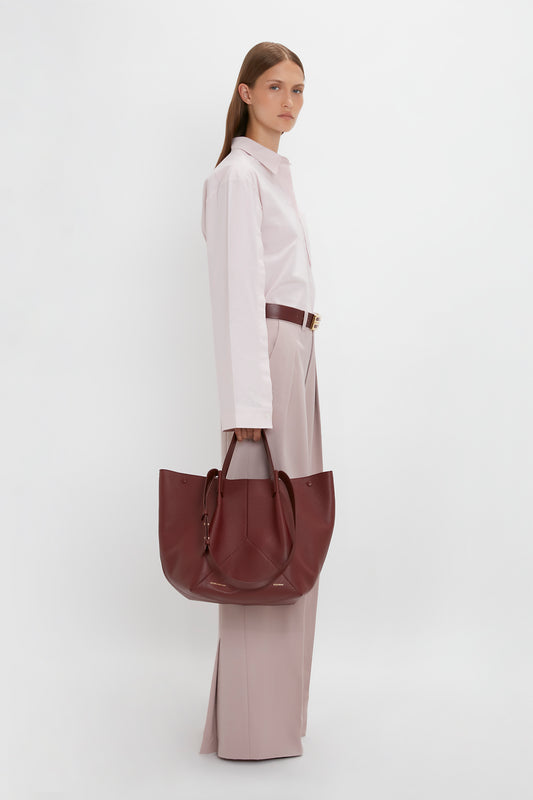 A person stands against a white background wearing a light pink buttoned shirt and light pink wide-legged trousers, holding a large maroon W11 Medium Tote Bag In Burgundy Leather by Victoria Beckham, perfect for versatile styling.