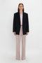 A woman stands against a plain white background wearing the Victoria Beckham Peak Lapel Jacket In Midnight over a light pink shirt and pale pink wide-leg trousers.