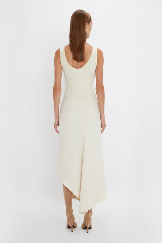 A person with long hair is standing with their back to the camera, wearing a Victoria Beckham Sleeveless Tie Detail Dress In Cream featuring an asymmetric hemline paired with nude heels.
